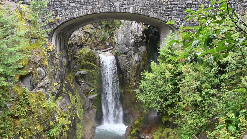 Stone bridge over narrow gorge with tall waterfall and lush green foliage in mountain forest setting