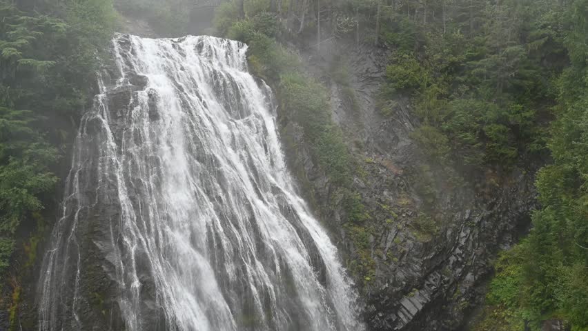Powerful waterfall plunges over rocky cliff surrounded by lush green forest in remote mountain wilderness
