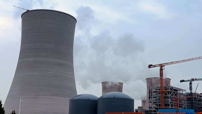 Massive power plant under construction, showing huge cooling towers emitting steam, industrial tanks, and a construction crane against a cloudy sky.