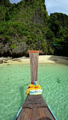 A longtail boat approaches a sandy beach in Phi Phi Islands, Thailand. Clear, shallow turquoise water surrounds the boat decorated with flower garlands. Suitable for travel marketing, tourism