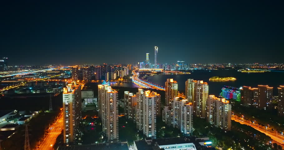 Aerial shot of the brightly illuminated modern cityscape with skyscrapers and a waterfront bridge at night in Suzhou, China.