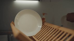 Man carefully placing clean white plate on wooden dish drying rack by kitchen sink finishing daily dishwashing task in contemporary home kitchen, close-up. Concept of dishwashing routine at home - Powered by Shutterstock - Get 15% off with code: PIKWIZARD15