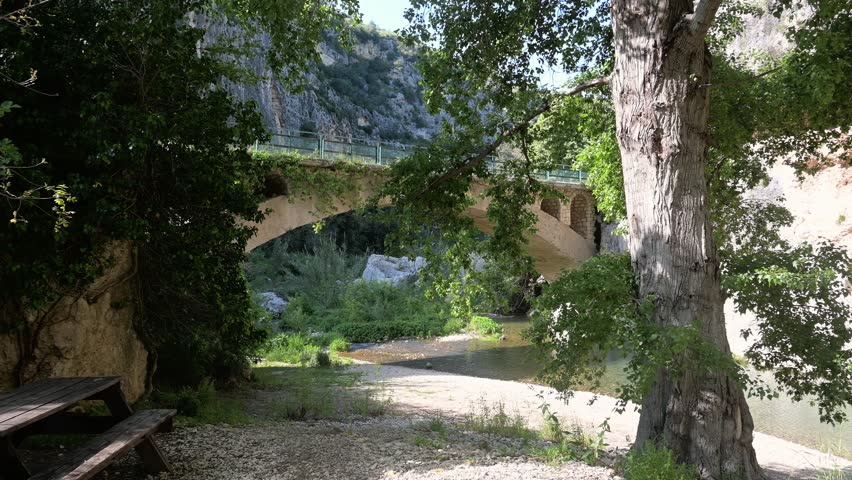 Panoramic view of a stone arch bridge crossing a calm river with cliffs, dense greenery, and shaded trees surrounding the gravel shore.