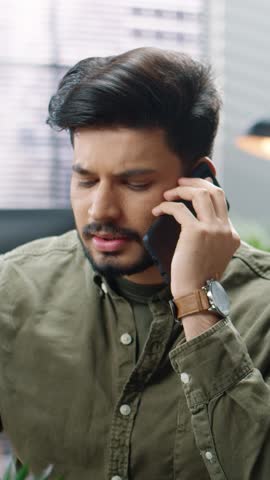 Serious male wearing olive green shirt holding phone to ear during conversation. Listening carefully and speaking calmly while managing business call in modern indoor office space.