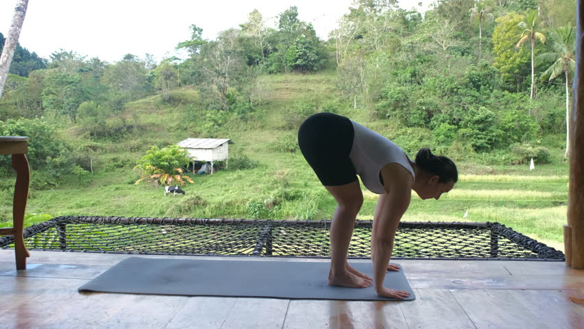 A young woman passionately engages in her yoga routine on a bamboo terrace, immersed in the beauty of tropical greenery and a peaceful rice plantation, slow motion