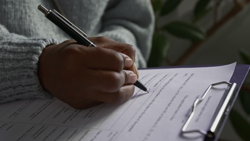 Close-up view of hand of unrecognisable Black young woman filling in visa application form holding clipboard and pen