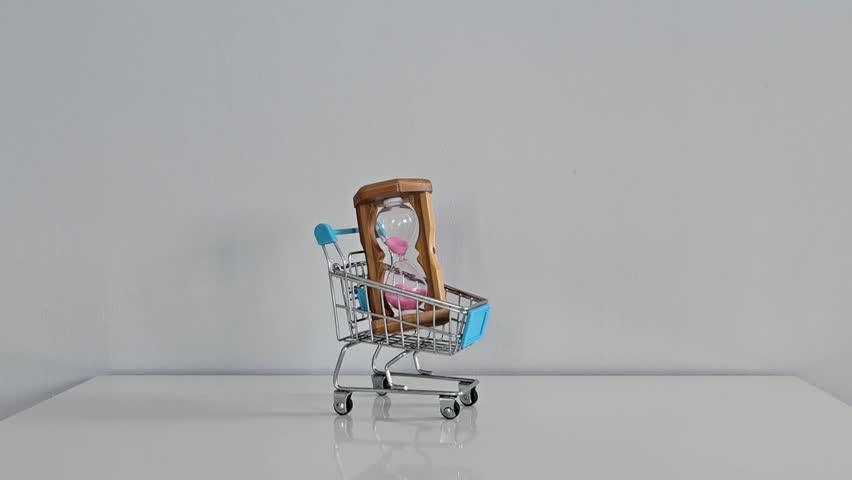 A hand picks up an almost empty hourglass, flips it, and places it back into a mini shopping cart on a white table with a gray background — symbolizing a fresh start or reset.