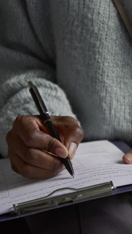 Vertical close-up view of hand of unknown Black young woman dressed in sweater filling in visa application form holding clipboard on her knee
