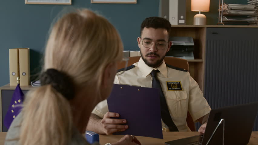 Over the shoulder view of young male visa processing officer dressed in uniform holding clipboard interviewing blonde senior woman at his workplace