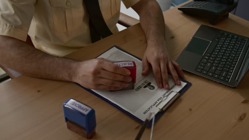 Unrecognisable male immigration officer in uniform stamping visa application of woman refused and giving clipboard to her while working at wooden desk with laptop and two stamps