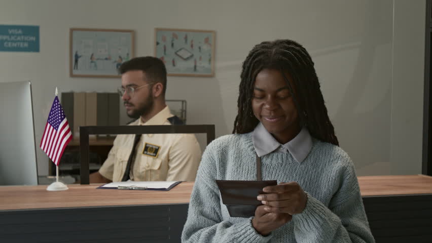 Portrait of happy African American girl showing passport with approved seal and smiling for camera in visa center, male officer working in background