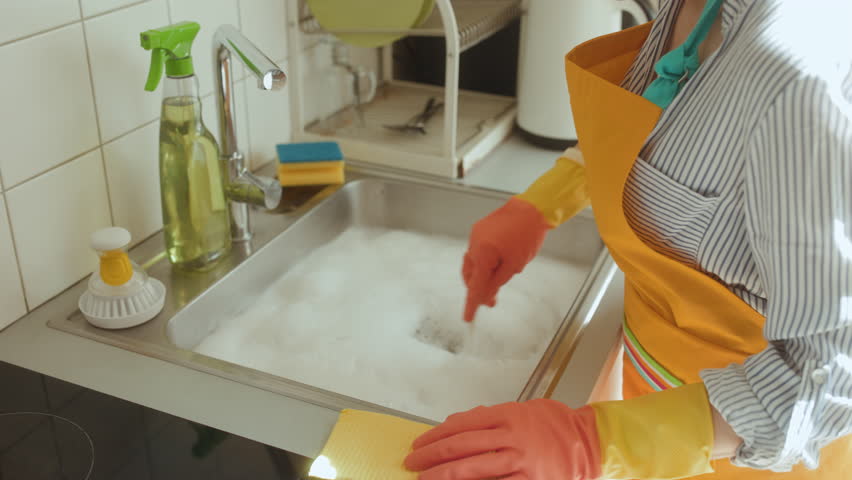A soapy sink hints at a clogged drain. Woman stirs the water, facing a common plumbing issue. Cleaning cloth and spray bottle nearby, seeking a solution.