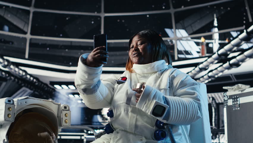 Cheerful female astronaut records selfie video with smartphone, speaking to subscribers during galactic mission. Jolly african american woman in starship recording content for followers, camera B