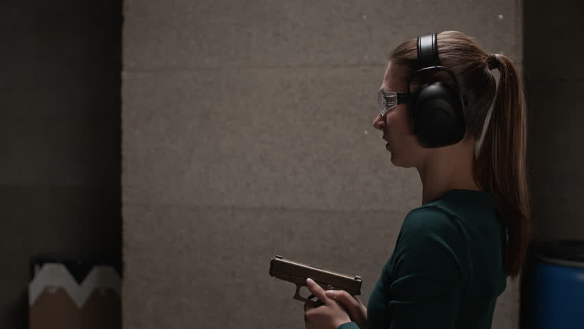 Side view of young Caucasian female trainee in ear protectors and goggles pointing and shooting pistol while practicing at indoor range