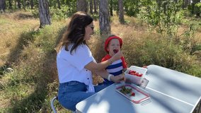 A woman is offering a strawberry to a child, who is laughing joyfully, as they sit at an outdoor table with containers of strawberries, surrounded by trees and dry grass on a bright, sunny day. - Powered by Shutterstock - Get 15% off with code: PIKWIZARD15