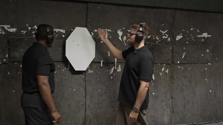 Medium long shot of Caucasian man discussing shot-up target sheet with male African American partner standing by wall in range