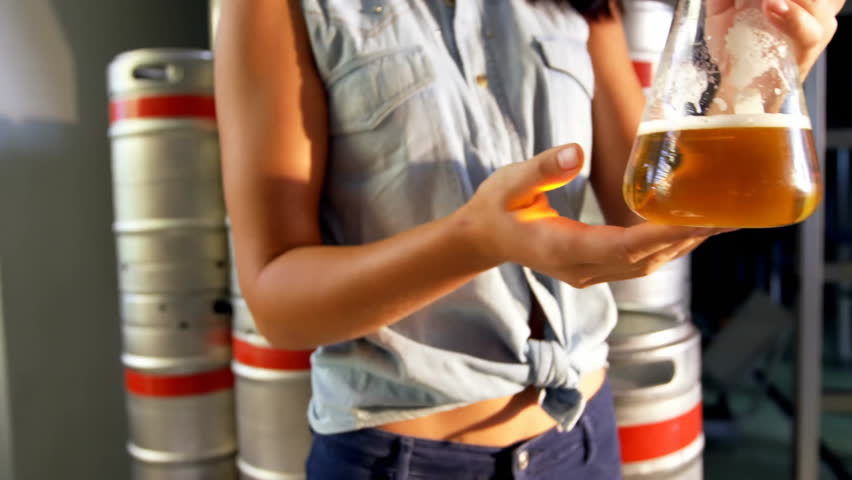 Female brewer lifting flask to light, rotating and inspecting sample amid holographic data charts. Laboratory, analytics, precision, industrial, innovation, technology, scientific