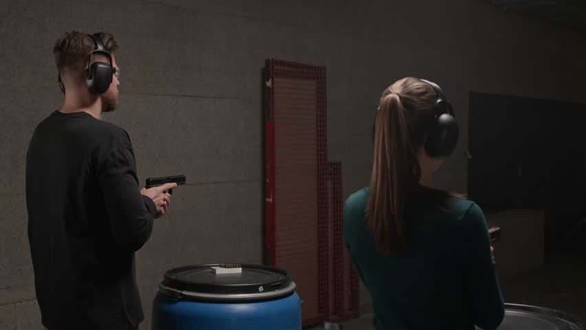 Back view of two people wearing earmuffs and goggles practicing shooting from guns at indoor range