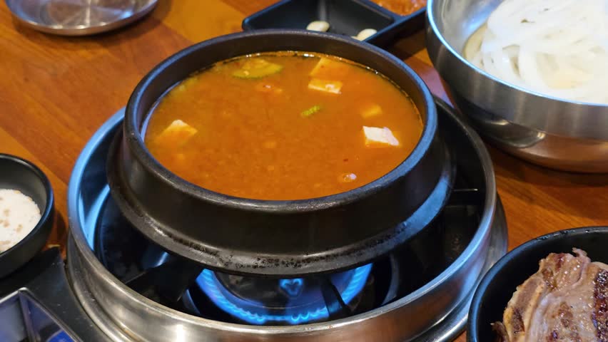 A close-up shot of a Korean traditional stew Doenjang jjigae - fermented soybean paste Korean cuisine stew with tofu seved in earthenware pot on a tabletop gas stove