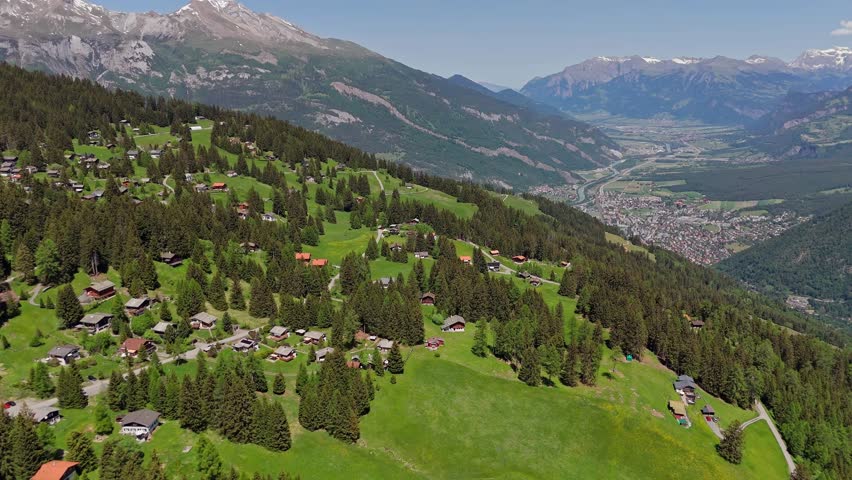 Idyllic swiss landscape with small neighborhood on green alp mountains in summer. Aerial approaching wide shot. Alp Panorama with snowy peaks.