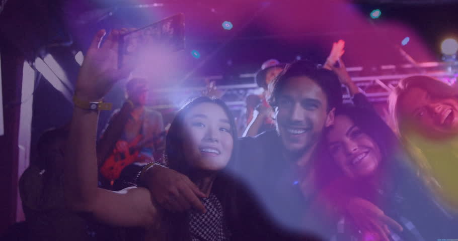 Friends extending selfie stick in club, capturing selfie amid animated colored lights for nightlife. Party, celebration, camaraderie, excitement, vibrant, nightlife, entertainment