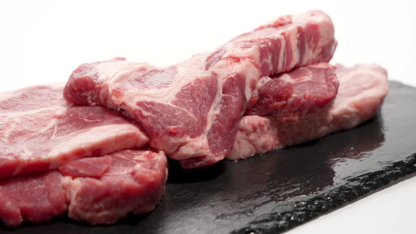 A stack of raw pork chops lies on a black stone slab. The rotating meat reveals juicy cut edges and marbling.