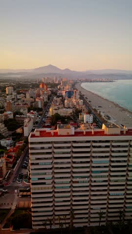 Forward-moving drone shot over the rooftops of Alicante’s coastal district at sunset. The image captures the expansive view of Muchavista beach, the Mediterranean Sea, residential buildings, palm-line