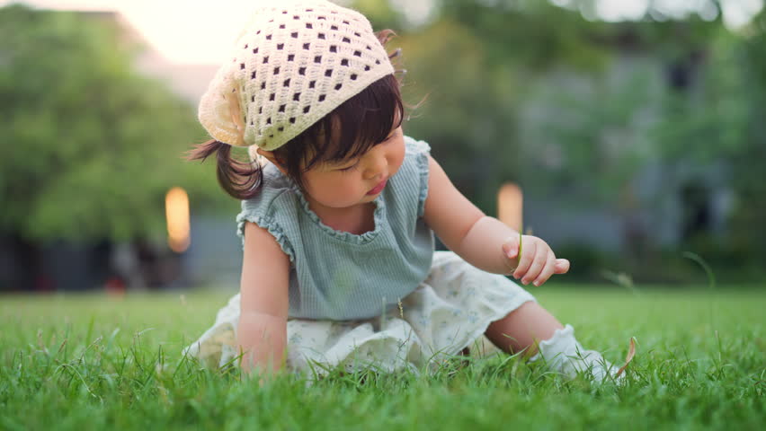 Happy little asian baby girl sitting on green grass lawn, touching and exploring nature in backyard garden during summer – learning, growth and outdoor fun