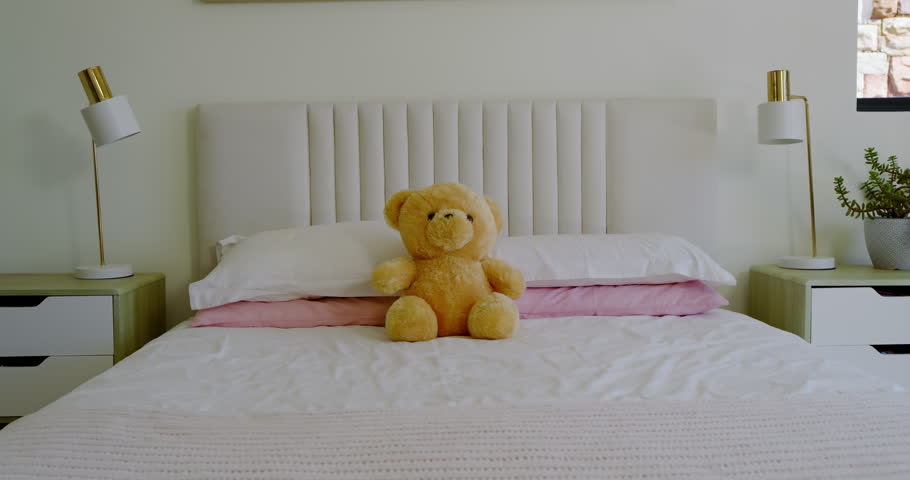 Camera is panning slowly right to frame teddy bear on bed between lamp, potted plant. Plush, serenity, minimalist, interior, decor, tranquility, comfort