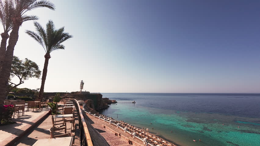 Establishing shot of sea resort beach with coral reefs, sunbeds, umbrellas, palm trees and lighthouse at background in the clear sunny morning. Red sea coastline in Sharm El Sheikh, Egypt