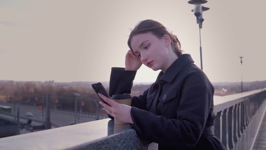 Casually dressed young woman relaxing with takeaway coffee, browsing smartphone while standing on pedestrian bridge, overlooking urban cityscape during golden sunset light
