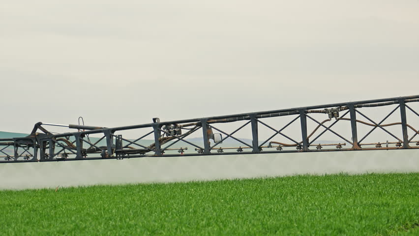 Agricultural sprayer boom in action over green field, Close-up view of nozzles dispersing mist evenly, Modern farming equipment for crop treatment