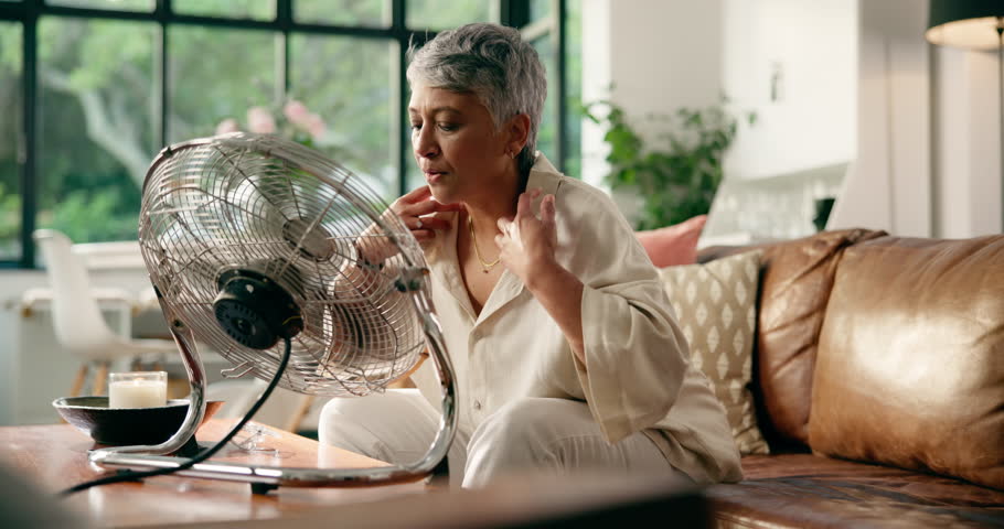 Senior woman, electric fan and cool down in home for hyperthermia and heat. Hot weather, propeller and person in living room for fresh air, ventilation or wind in summer for menopause in retirement