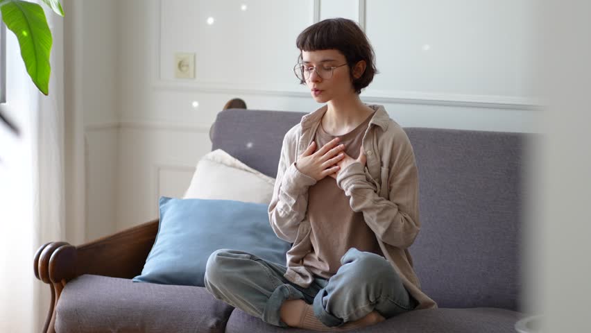 Young woman meditating with deep breaths hands on chest sit in lotus position to calm mind at home. Mindfulness stress relief, spiritual controlled breathing, inner emotional regulation, zen routine