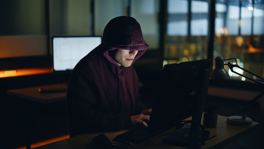 Hooded hacker breaking data servers at night room closeup. Dangerous man getting access making cyber attack in corporate center place. Spy cracker infecting system with virus alone at dark atmosphere 