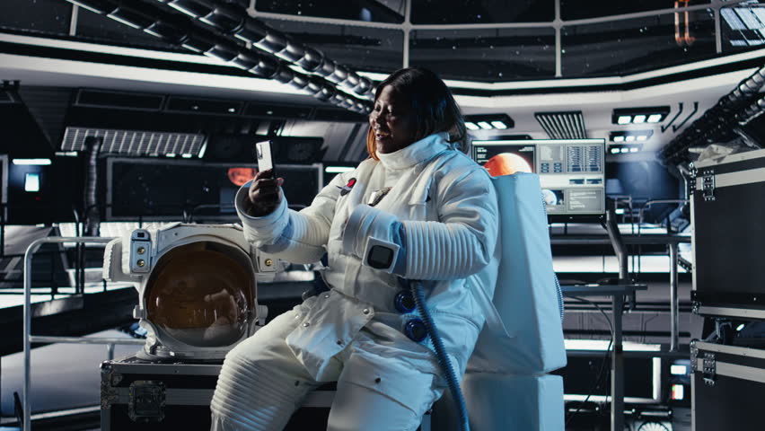 Cheerful female astronaut records selfie video with smartphone, speaking to subscribers during galactic mission. Jolly african american woman in starship recording content for followers, camera A