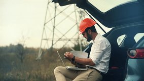 Engineer in orange hard hat working on a laptop and clipboard from the back of a car near power lines. Fieldwork and remote technical inspection in an industrial or energy-related environment survey - Powered by Shutterstock - Get 15% off with code: PIKWIZARD15