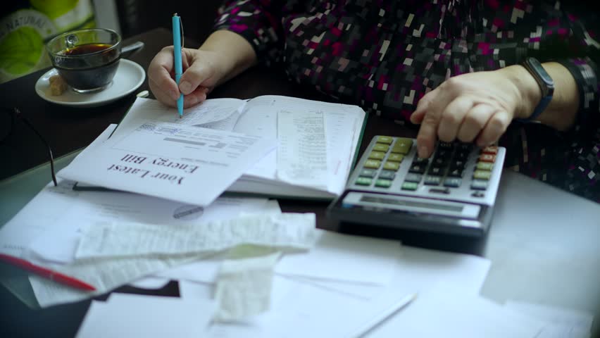 Senior woman calculating home finances using a large calculator, pen, and documents. Energy bills, receipts, and notebook on desk. Personal budget and expense management. Utility Bills tax calculation - Powered by Shutterstock - Get 15% off with code: PIKWIZARD15