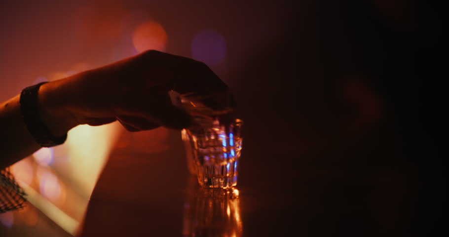 Nightclub Bartender Puts Filled Shot Glasses on a Bar Table. Close Up Footage with a Hand Moving Shots in a Moody, Colorful Party Environment. People on the Other Side Waiting for Their Spirits