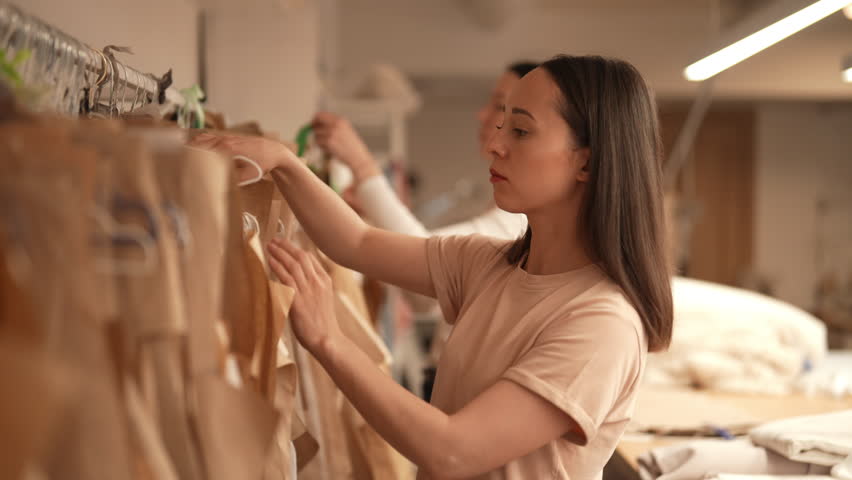 Portrait of professional seamstress choosing sewing pattern hanging on clothes rack in bright atelier, preparing to create new clothing collection. Concept of sewing manufacturing, slow motion.