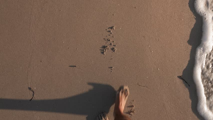 POV shot capturing a man’s barefoot footsteps walking along a sandy coastline. The camera follows the steps, showcasing the texture of the sand and the gentle lapping of waves in the background.
