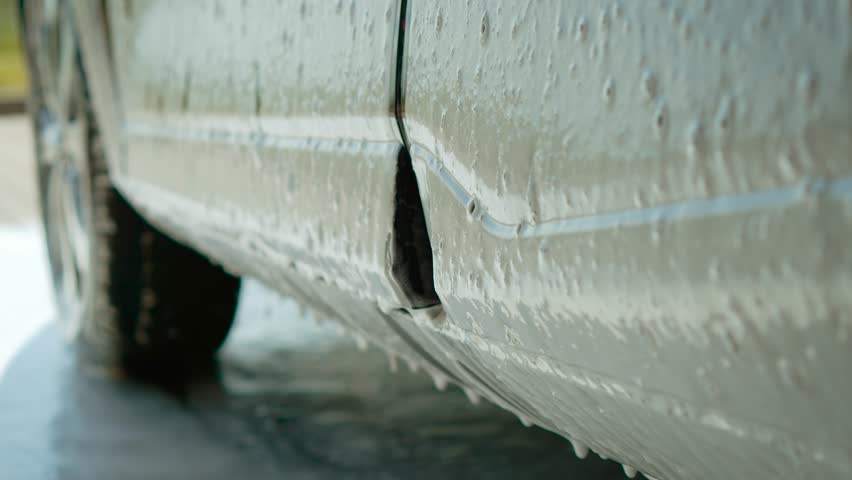 Close-up of foam dripping from lower car door, drops falling, self-service wash with active foam. Soap flowing down side panel, suds dripping, foamy car wash station. Thick lather sliding from door
