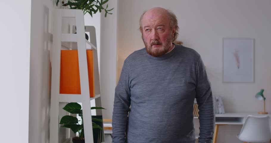 Tired senior man clutching his head and leaning on wall while suffering from dizziness at home. Exhausted weary elderly male having vertigo, headache or blood pressure increase, losing consciousness.