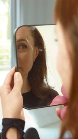 Woman with injection marks on facial skin looking at reflection in mirror, vertical orientation. Client examining post treatment face after aesthetic procedure. Patient checking results of