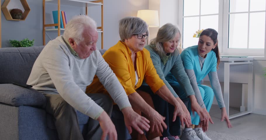 Group of happy senior elderly people sitting on sofa together with nurse watching TV and doing sport exercises in retirement home. Leisure in nursing home concept. 4k video. Slow motion video.