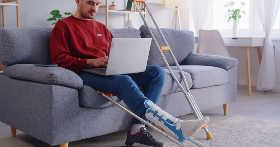 Injured man with leg in plaster cast sitting on sofa with crutches nearby, talking on smartphone and working on laptop. Young man engaged in remote work during recovery from foot trauma at home.