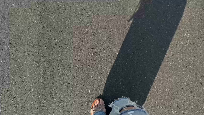 Top view of bare feet in sandals walking on dark asphalt in ripped jeans on a sunny day, showing casual summer movement and relaxed urban lifestyle
