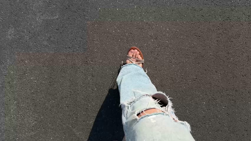 Top view of bare feet in sandals walking on dark asphalt in ripped jeans on a sunny day, showing casual summer movement and relaxed urban lifestyle