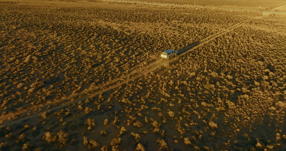 Drone shot of a silver truck driving on a dirt road though the Nevada high desert at sunset. Long shadows stretch over the sage brush. Camera flies over and pans left