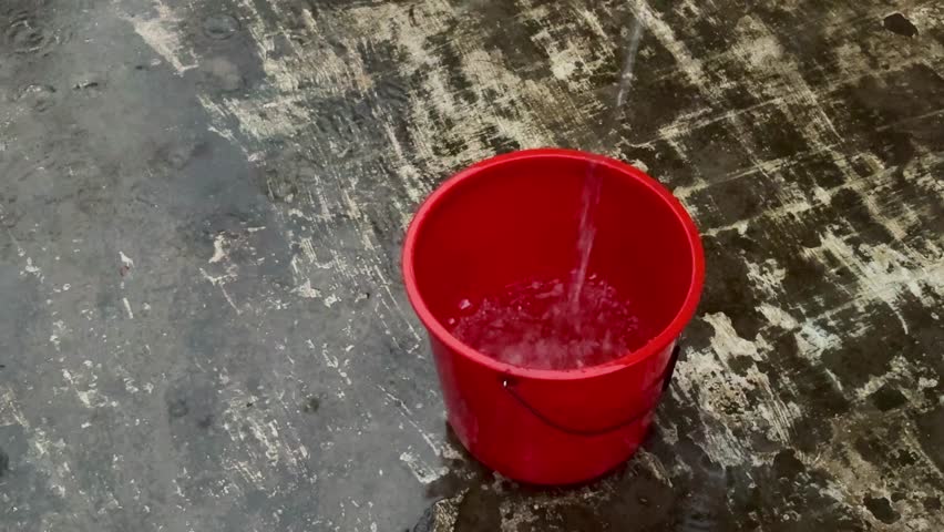 A red bucket on a cement floor collecting rainwater 